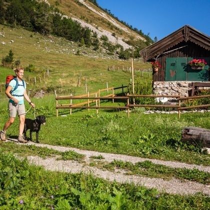 Wanderin mit angeleintem Hund neben einer Alm an einem Weidezaun